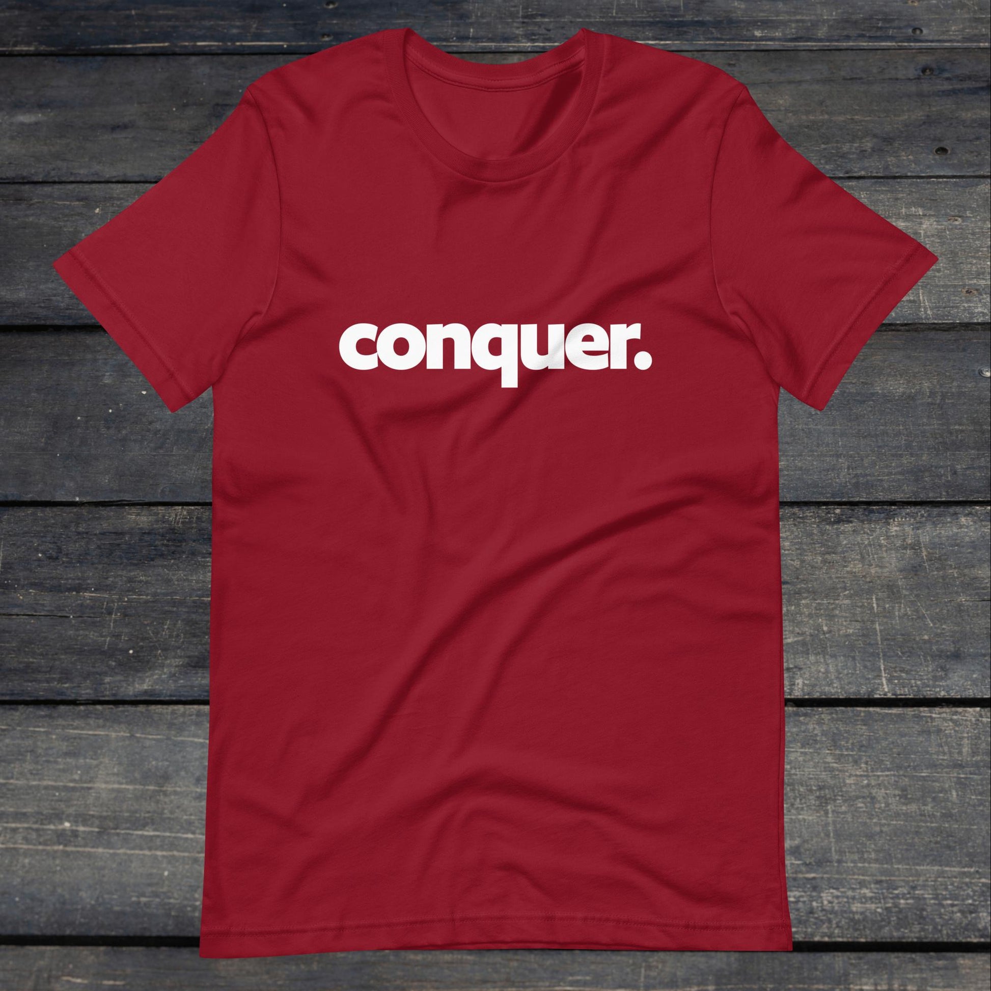 A photo of the Conquer Tee. This tee is cardinal red with white text that reads conquer in large bold lettering.