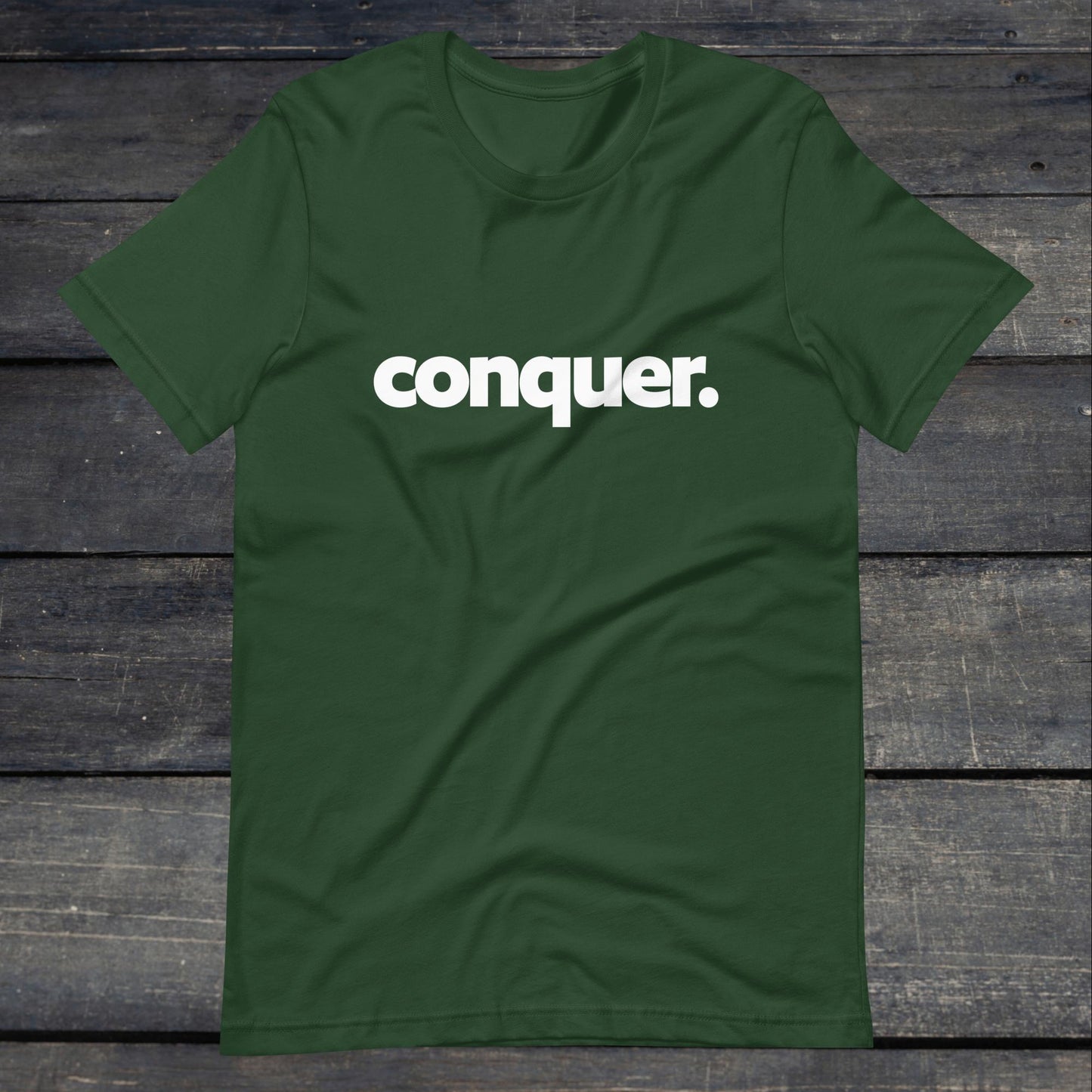 A photo of the Conquer Tee. This tee is forest green with white text that reads conquer in large bold lettering.