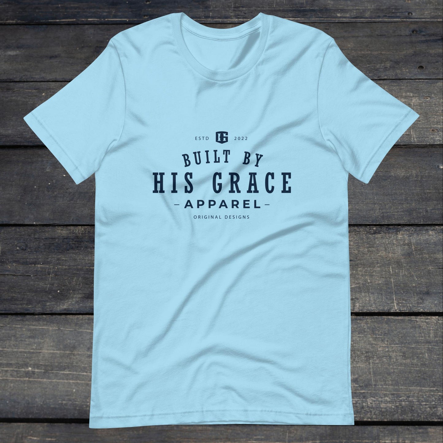 A photo of the "Built By His Grace Apparel" Tee. This tee is ocean blue with navy blue text that reads "Built By His Grace Apparel". 