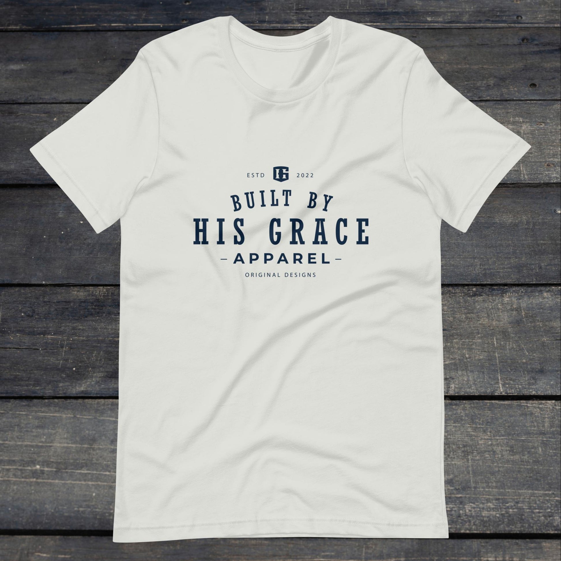 A photo of the "Built By His Grace Apparel" Tee. This tee is silver with navy blue text that reads "Built By His Grace Apparel". 