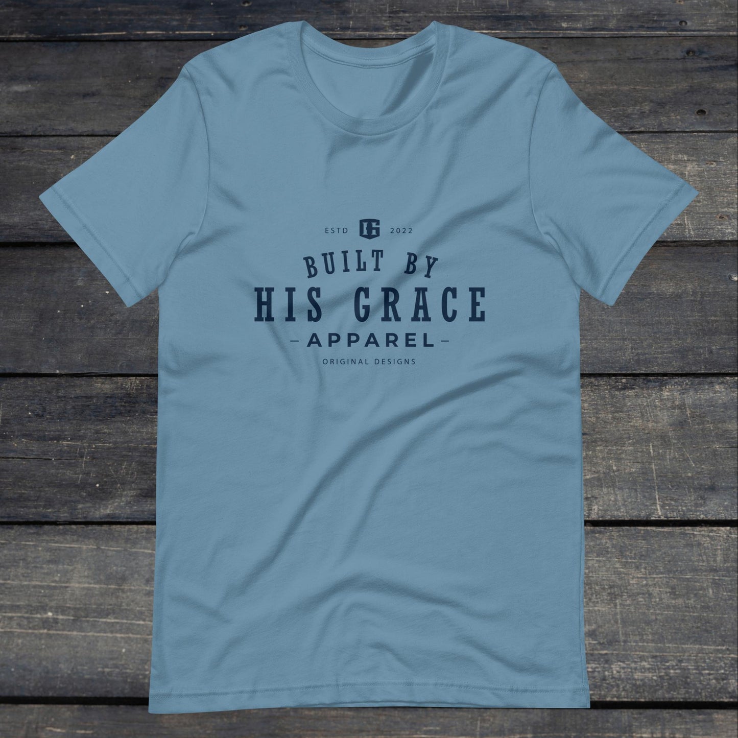 A photo of the "Built By His Grace Apparel" Tee. This tee is steel blue with navy blue text that reads "Built By His Grace Apparel". 