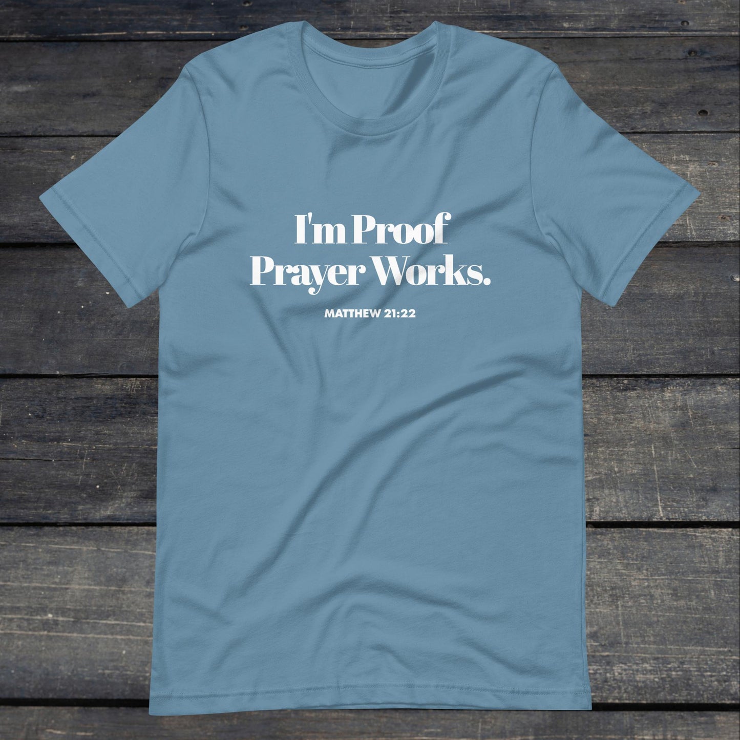 A photo of the "I'm proof prayer works" tee. The shirt color is steel blue with white text that reads "I'm proof prayer works".